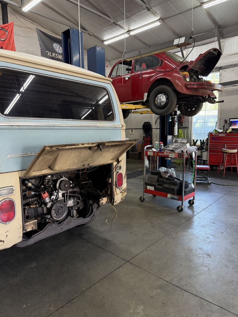 Bay-window Volkswagen Bus with the engine lid open and a red Beetle on the lift behind it